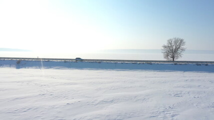 Aerial shot of white car riding through snow covered road near a frozen lake. SUV driving at countryside route near icy pond at sunny winter day. Flying over the auto moving through scenic landscape