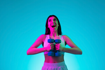 Laughting. Brunette woman's portrait on blue studio background in mixed neon. Beautiful model training with dumbbells inspired. Concept of human emotions, facial expression, sales, ad, fashion, sport.