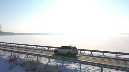 Aerial view of car riding through snow covered road near frozen lake. White SUV driving at dam route on winter day. Fly over the auto moving through bridge of river. Scenic landscape way. Drone shot