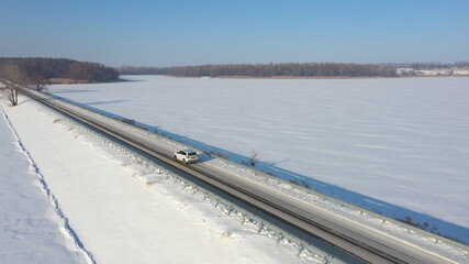 Aerial shot of car riding through snow covered road near frozen lake. White SUV driving at dam route on winter day. Flying over the auto moving through bridge of river. Scenic landscape way. Top view
