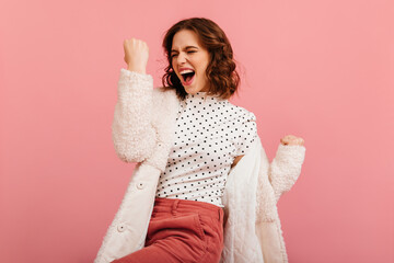 Excited curly woman raising fist. Studio shot of happy girl in coat isolated on pink background.