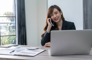 An Asian female finance worker sits on the phone with a customer who calls in to consult. At the office table, sit and work happily and smiling, concept over the phone.