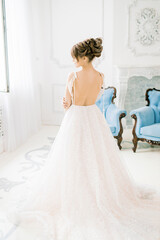 Fototapeta premium Beautiful girl the bride stands with an open back in a wedding dress. Boudoir and morning of the bride. Dress with tassels. In a white interior with blue classic chairs.