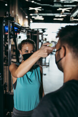 Young female fitness gym worker and instructor measuring body temperature of young sporty man with infrared thermometer. Coronavirus world pandemic and sport theme.