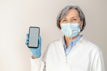 Fototapeta na wymiar Senior female doctor in a medical robe, protective face mask, and surgeon latex gloves posing isolated over gray background, demonstrating a smartphone with a blank screen for advertisement