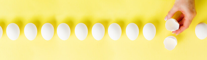 Fototapeta na wymiar White raw chicken eggs lying in row on a yellow trendy background and female hand holds broken egg. Easter minimal creative composition. Spring Happy Easter holiday card. Top view.