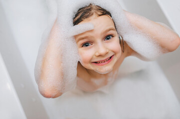 A small, red-haired girl, a child washes, sitting in a white bath with foam from soap, shampoo and...