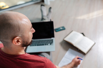 The concept of online courses and online learning. A bald man is working at a laptop and writing something down on paper. Rear and top view, close-up