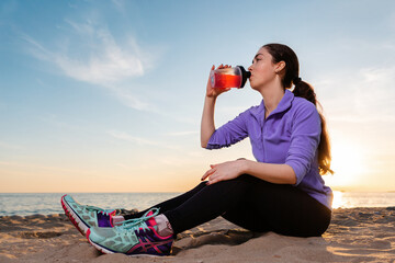 A young woman sits on the sand and drinks a protein shake from a shaker. Bottom view. The concept...