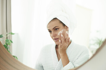 Beautiful young woman applying cream in bathroom. Face cream. Skin care. Beautiful smiling woman with a towel on her head after showering