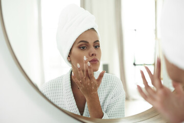 A beautiful young woman with vitiligo on her hands in a bathrobe and with a towel on her head applyies a cream for problem skin to her face. The concept of skin care, moisturizing and cleansing the