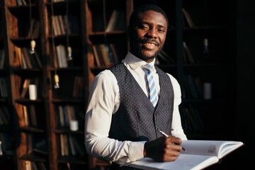 The teacher holds a notebook with a pen in his hands among the shelves with books