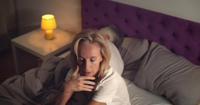 Senior woman sitting in bed and feeling stressed with husband sleeping on background