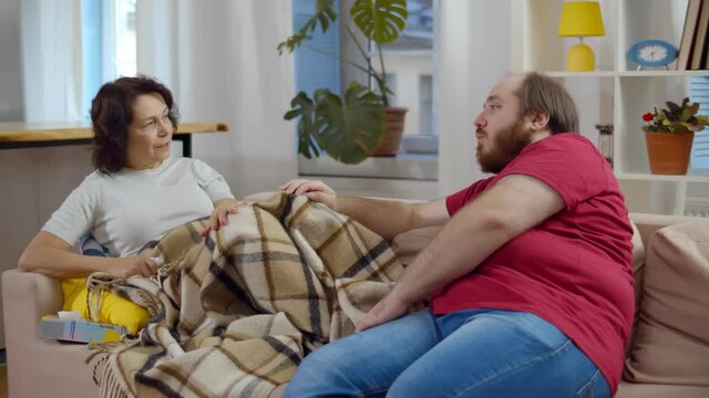 Young overweight man taking care of his sick mom sitting together on couch