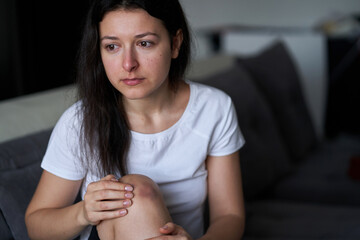Portrait of beaten woman with tears of her face and bruise on knee sit on the sofa