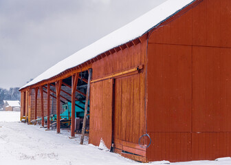 A photograph of a very old red barn with a turquoise tractor inside. The barn roof is covered in snow. 