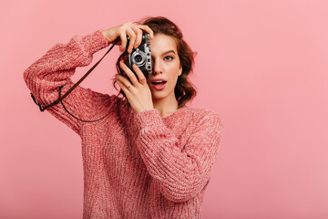 Surprised girl in knitted sweater taking photos. Studio shot of female photographer holding camera on pink background. - Powered by Adobe