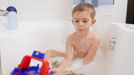 Cute little boy taking bath at home and playing iwth colorful toy ship or boat. Concept of child hygiene and health care at home. Kids having fun and playing