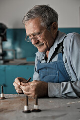 An elderly master with glasses at work. A white old man of Caucasian appearance in work clothes sits at a table and works with his hands.