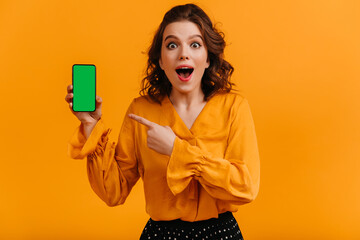Naklejka premium Excited woman pointing with finger at digital device. Studio shot of shocked girl holding smartphone with blank screen.