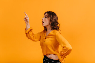 Surprised caucasian woman looking up on yellow background. Studio shot of shocked well-dressed girl pointing with finger.