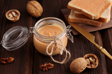 Fototapeta na wymiar Natural nut butter in a glass jar, a golden knife, walnuts and sandwiches with toast on a wooden brown background. Vegetarian traditional breakfast close up.
