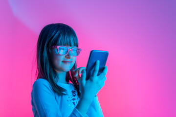 Happy fashionable young girl with a beauty smile holding a smart phone and typing a message on a multicolored neon pink light in the studio.