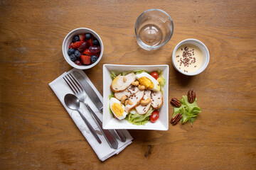 Top view of healthy and nutritious food, chicken salad, lettuce and tomatoes, bowls with fruits: strawberry, grapes and blueberries and another with dressing, a glass and cutlery on a cloth napkin