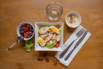 Top view, chicken salad with lettuce, egg, cherry tomatoes, peanuts and flaxseed, bowls one with dressing and another fruit strawberries, grapes, blue berry, a glass, knife and fork on a cloth napkin