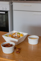 Chicken salad with lettuce, egg, peanuts and flaxseed in a white square bowl, small bowls one with dressing and one with walnuts on a wooden table, blurred background. Healthy and nutritious food