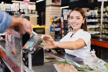 Smiling cashier looking at camera near customer paying with smartphone on blurred foreground in supermarket