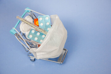 Shopping cart and medicine. An expensive medicine. Cough pills shopping basket with thermometer and medicine mask on blue background.