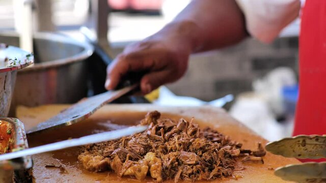 Close up of a Mexican food stand, where a serving of juicy birria (stewed beef) is being chopped with a large butcher knife.  The steaming meat is tossed with prongs as it is cut up.
