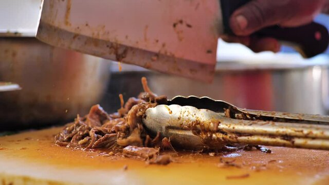 Close up of a Mexican food stand, where a serving of birria (stewed beef) is being chopped with a large butcher knife.  The steaming meat is tossed with prongs as it is cut up.
