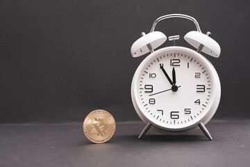  classic white alarm clock stands next to a bitcoin coin and the time shows 5 minutes to 12