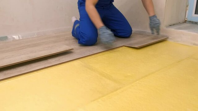 A man in a blue uniform and gloves repairs the floor of a room. The process of laying laminate on a yellow substrate. Floor repair