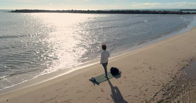 mujer madura haciendo postura loto en la playa frente al sol y dron a su espalda