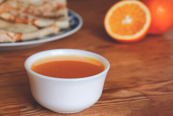 Orange syrup in a white bowl and oranges and pancakes in the background.