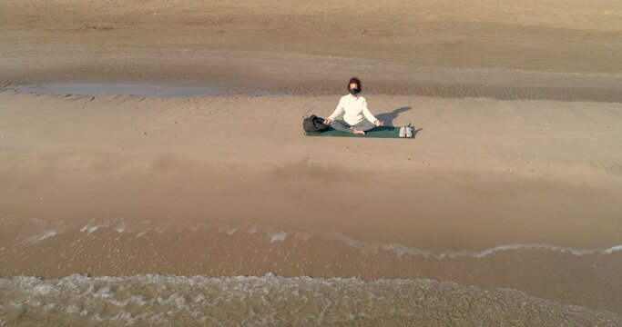 Dron se aleja de mujer sentada meditando en esterilla al borde del mar
