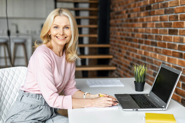 Enchanting middle-aged businesswoman looks at camera and smiles. Close-up portrait of charming blonde woman, female ceo sitting at the desk with a laptop in the loft style office
