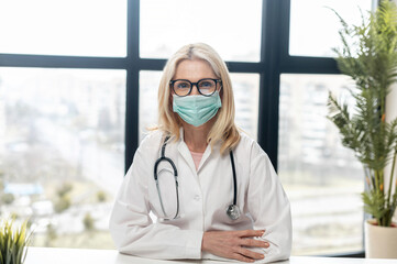Close-up portrait of confident female MD, physician, therapist, pediatric in protective face mask standing with arms crossed and looks at camera in modern hospital office. Healthcare and medicine