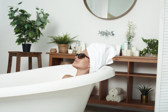 The pathos beautiful girl with vitiligo lies in the bath in the cat's sunglasses and a towel on her head. The concept of fashion, skin care and style