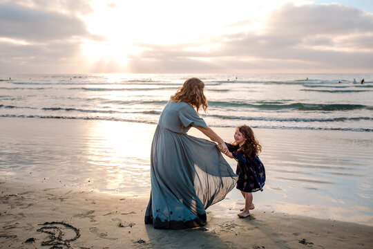 Mom in long dress playing with 3 yr old daughter on beach at sunset