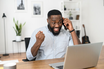 Obraz premium Successful Afro American freelancer talking on the phone, happy to achieve the goal. Young businessman looking at the screen of the laptop, winner, receiving good news in successful conversation