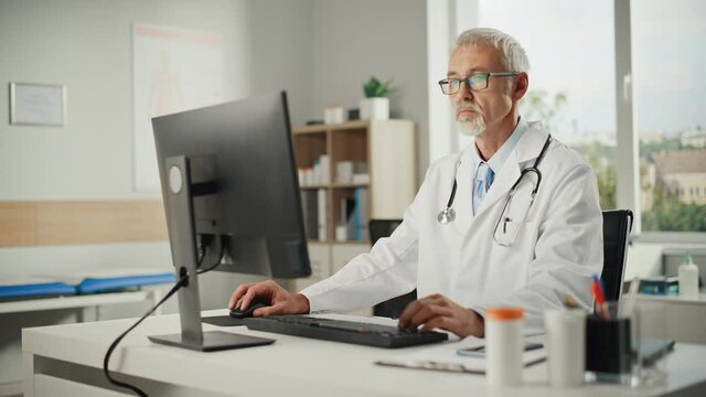 Experienced Middle Aged Male Doctor Wearing White Coat Working on Personal Computer at His Office. Senior Medical Health Care Professional Working with Test Results, Patient Treatment Planning.