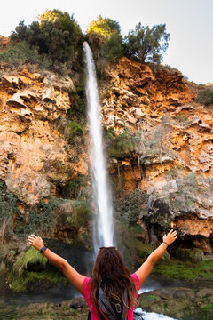 Rear view of a young woman with a backpack with open arms in front of a large waterfall