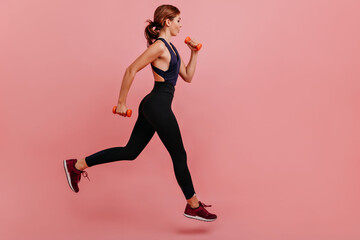 Fototapeta na wymiar Strong hardy woman trains on pink isolated background. Girl in black leggings runs with weight in her hands