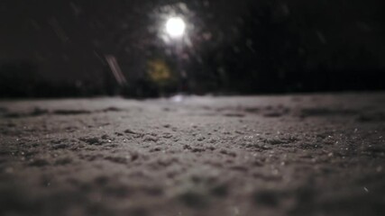 Low angle pan shot of snow-covered ground during snowflakes falling down from sky at night. Blurred lantern light in background.