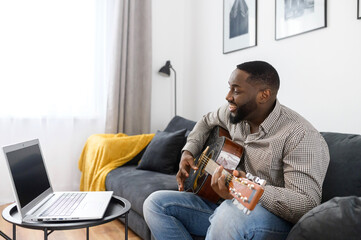 Smiling young African American male musician playing guitar during an online concert at home while...