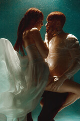 The couple is dancing or hugging in the pool underwater. A girl in a dress with a long train and a guy swim underwater